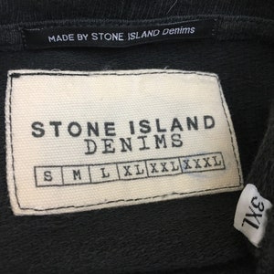 May include: Close-up of a black garment tag with the text "MADE BY STONE ISLAND Denims". Below is a white tag with "STONE ISLAND DENIMS" and size options: S, M, L, XL, XXL, XXXL. A smaller tag reads "3XL".