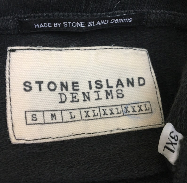 May include: Close-up of a black garment tag with the text "MADE BY STONE ISLAND Denims". Below is a white tag with "STONE ISLAND DENIMS" and size options: S, M, L, XL, XXL, XXXL. A smaller tag reads "3XL".