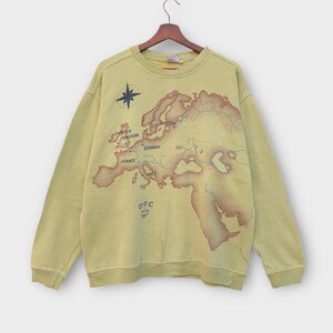 Puede incluir: Sudadera amarilla clara con un diseño de mapa vintage de Europa y África del Norte. El mapa incluye nombres de países y una rosa de los vientos. El texto "OTC MCM" está impreso en la parte inferior izquierda.