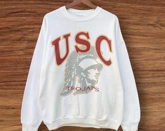 May include: A white crewneck sweatshirt with the letters "USC" in maroon and gold, above a silver Trojan warrior graphic. The word "TROJANS" is printed in maroon below the graphic. The sweatshirt is hanging on a wooden hanger.