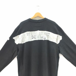 May include: Black sweatshirt with the text "STONE ISLAND DENIMS" printed across the back in a distressed, white font. The sweatshirt is hanging on a wooden hanger against a white background.