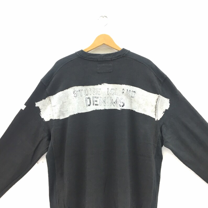 May include: Black sweatshirt with the text "STONE ISLAND DENIMS" printed across the back in a distressed, white font. The sweatshirt is hanging on a wooden hanger against a white background.