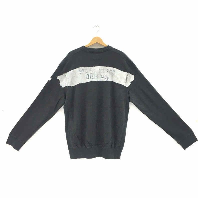 May include: Black sweatshirt with long sleeves, featuring a distressed gray horizontal stripe across the back with the text "STONE ISLAND DENIMS". The sweatshirt is displayed on a wooden hanger against a white background.