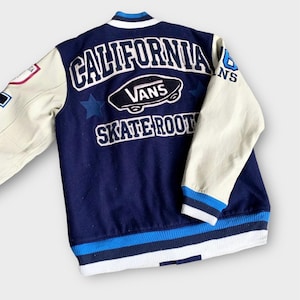 Pode incluir: Uma jaqueta de beisebol azul e branca com "CALIFORNIA" e "VANS SKATE ROOTS" bordados nas costas. As mangas são brancas com listras azuis.
