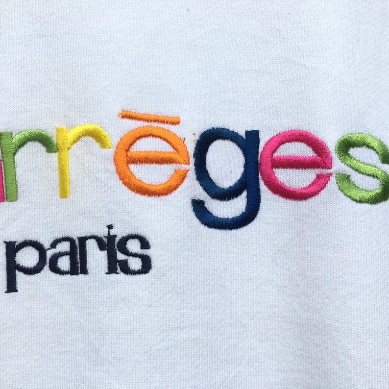 May include: White fabric featuring embroidered text in various colours. The word "arrēges" is embroidered in green, yellow, orange, blue, pink, and green. Below, the word "paris" is embroidered in navy blue.