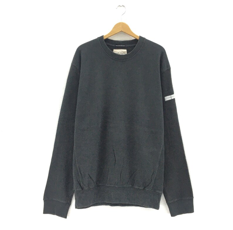 May include: A dark gray sweatshirt with a crew neck and long sleeves, hanging on a wooden hanger. The sweatshirt has a small tag on the upper chest and a reflective stripe detail on the left sleeve. The fabric appears to be a soft, comfortable material.