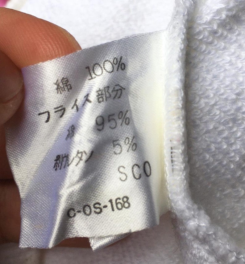 May include: A close-up of a white fabric label with Japanese text and percentages. The label states "100% cotton" and "95%" with other text and numbers. The label is attached to a white garment.