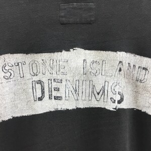 May include: Close-up of a black sweatshirt with a white, distressed graphic. The graphic reads "STONE ISLAND DENIMS" in a stencil font. The sweatshirt has a small, rectangular patch near the neckline.