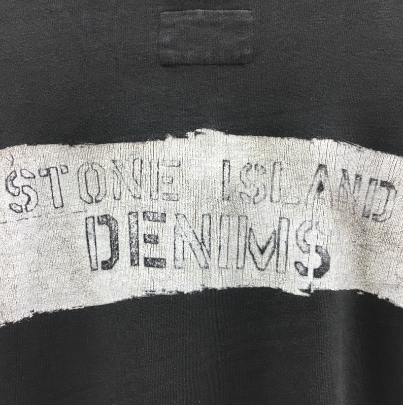 May include: Close-up of a black sweatshirt with a white, distressed graphic. The graphic reads "STONE ISLAND DENIMS" in a stencil font. The sweatshirt has a small, rectangular patch near the neckline.