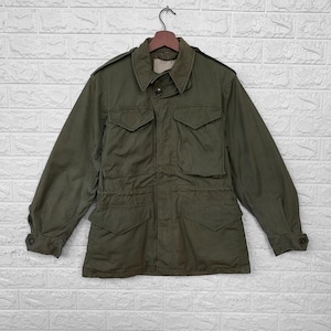 May include: Olive green military-style jacket with multiple front pockets and button closures. The jacket has a collared neckline and long sleeves with buttoned cuffs. It is hanging on a wooden hanger against a white brick wall.