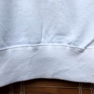May include: Close-up of a white sweatshirt. The image focuses on the ribbed hem and the lower portion of the garment. The fabric appears soft and slightly textured, with subtle folds and shadows.