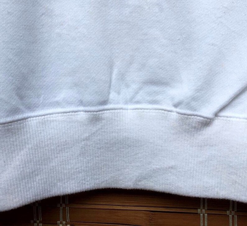 May include: Close-up of a white sweatshirt. The image focuses on the ribbed hem and the lower portion of the garment. The fabric appears soft and slightly textured, with subtle folds and shadows.