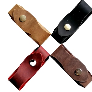 Könnte beinhalten: Vier Gitarrenplektrumhalter aus Leder in verschiedenen Farben: Hellbraun, Schwarz, Rot und Braun. Jeder Halter hat einen Messing-Druckknopfverschluss. Die Halter sind kreuzförmig auf weißem Hintergrund angeordnet.