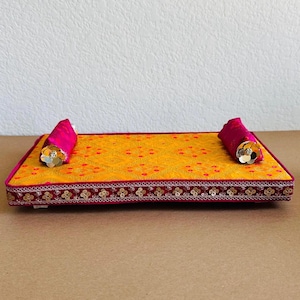 May include: A decorative rectangular tray with a yellow fabric top and a red dot pattern. It has a magenta border with gold embellishments and two magenta bolster pillows with silver flower accents.