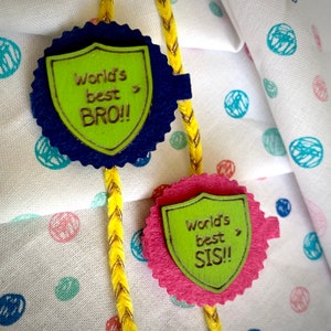 May include: Two decorative friendship bracelets with the text "World's best BRO!!" and "World's best SIS!!". The medallions are green and blue, and pink and green, with a yellow and gold braided cord.