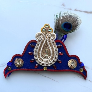 May include: A blue and red crown with a white and gold beaded design and a peacock feather accent.