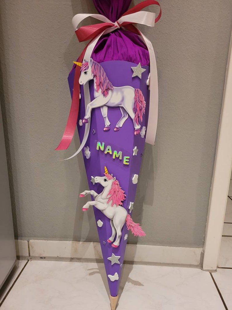 May include: Purple and pink school cone with a unicorn design. The cone has a white and red ribbon tied around it. The cone has the word "NAME" written on it.