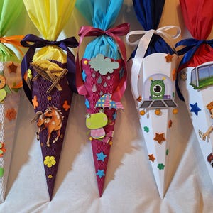 Könnte beinhalten: Fünf bunte Papierkegel, die mit verschiedenen Themen verziert sind. Die Kegel sind mit Eulen, Pferden, Regenwolken, Außerirdischen und Fußballspielern verziert. Die Kegel sind mit Bändern gebunden und bereit für einen besonderen Anlass.