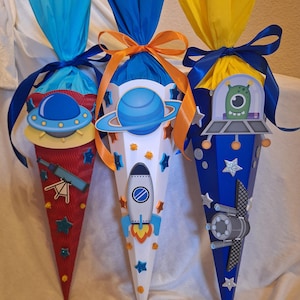 Könnte beinhalten: Drei farbenfrohe, kegelförmige Geschenktüten mit Weltraum-Dekorationen. Eine ist rot mit Teleskop und UFO, eine andere weiß mit Rakete und die dritte blau mit Alien und Raumschiff. Jede ist mit Seidenpapier und Bändern versehen.