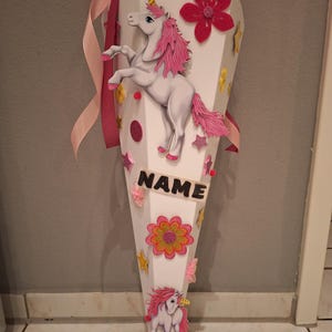 May include: A white cone-shaped school bag decorated with pink and gold glitter stars, flowers, and a unicorn. The bag has a pink ribbon and the word "NAME" written on it.