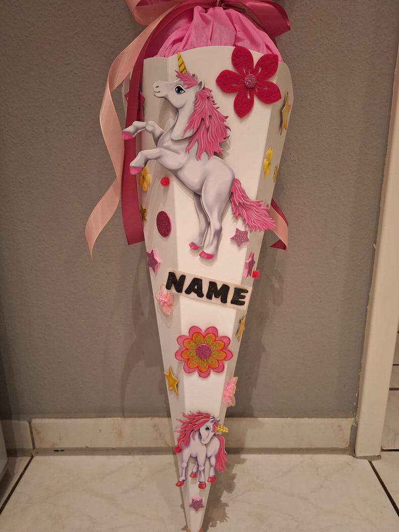 May include: A white cone-shaped school bag decorated with pink and gold glitter stars, flowers, and a unicorn. The bag has a pink ribbon and the word "NAME" written on it.