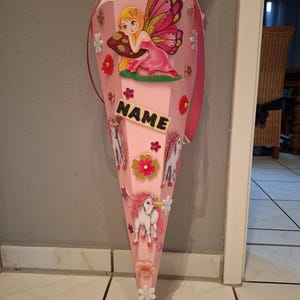 May include: Pink school cone decorated with a fairy, unicorns, and flowers. The cone has a tag that says "NAME".