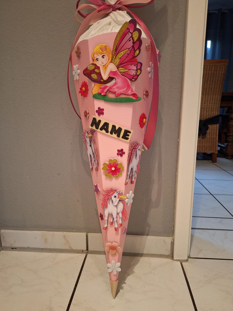 May include: Pink school cone decorated with a fairy, unicorns, and flowers. The cone has a tag that says "NAME".