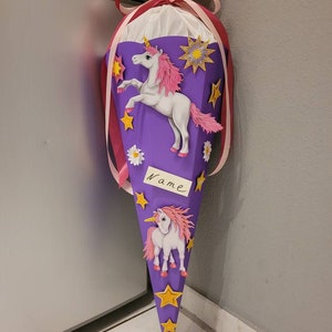 May include: Purple school cone with a unicorn design. The cone has yellow stars, pink flowers, and two white unicorns with pink manes. The cone has a white tag with the word "Name" written on it.
