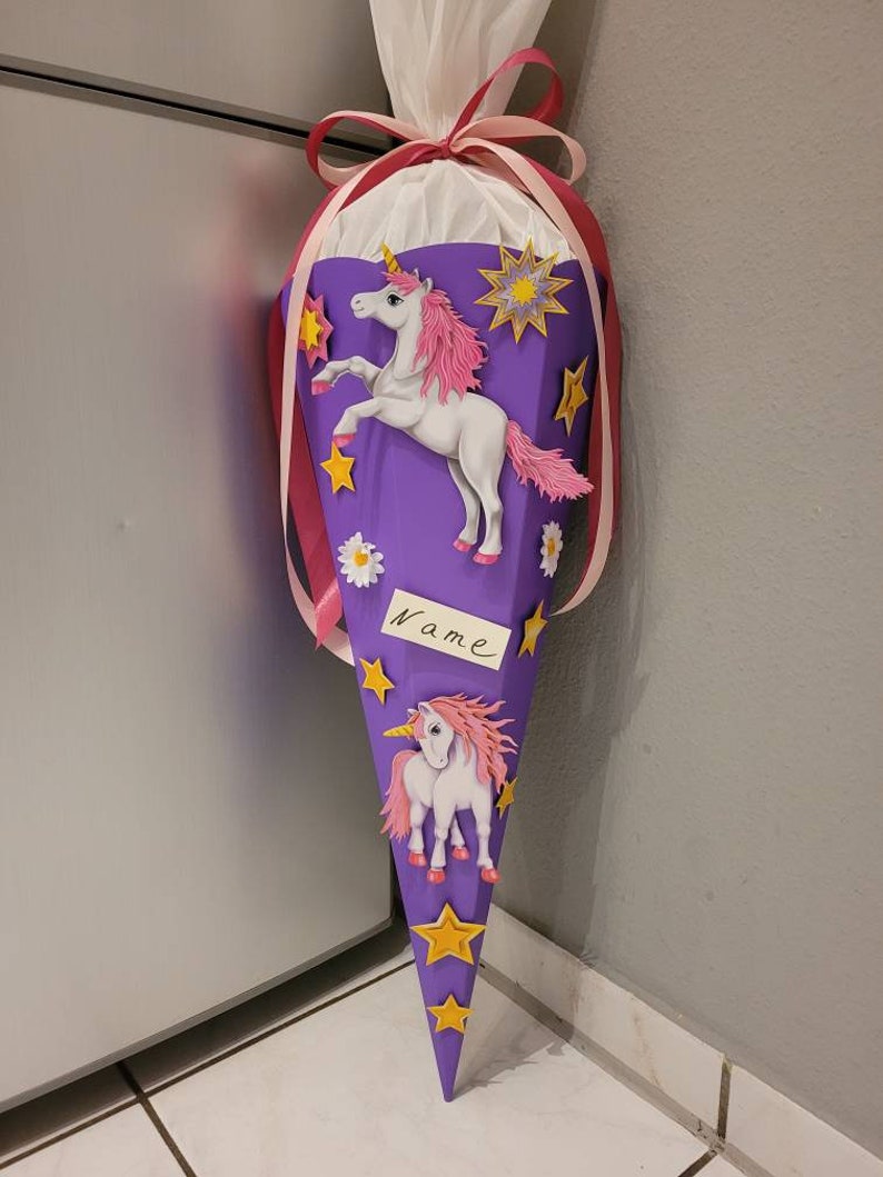 May include: Purple school cone with a unicorn design. The cone has yellow stars, pink flowers, and two white unicorns with pink manes. The cone has a white tag with the word "Name" written on it.