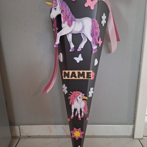 May include: A black and pink school cone with a unicorn design. The cone has a pink ribbon and a name tag that says "NAME".