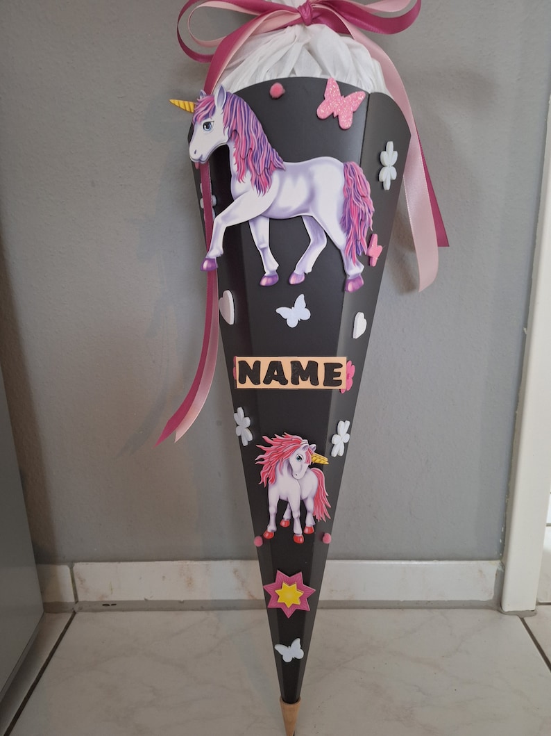 May include: A black and pink school cone with a unicorn design. The cone has a pink ribbon and a name tag that says "NAME".