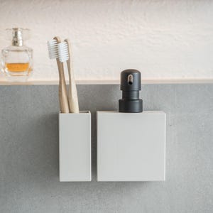 May include: Two white toothbrushes in a white ceramic holder next to a white ceramic soap dispenser with a black pump.