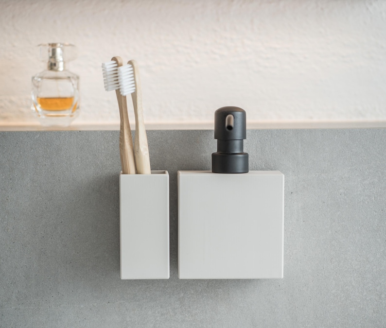 May include: Two white toothbrushes in a white ceramic holder next to a white ceramic soap dispenser with a black pump.