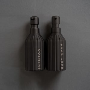 Może przedstawiać: Dwie czarne, żebrowane butelki z pompkami, jedna z etykietą "SZAMPON", a druga "PŁYN DO KĄPIELI".