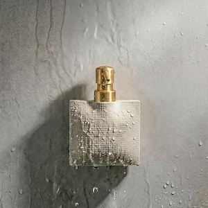 Op de afbeelding: Een vierkante, getextureerde, witte zeepdispenser met een goudkleurige pomp. De dispenser is nat met waterdruppels, wat duidt op recent gebruik. De dispenser is op een grijze muur gemonteerd.