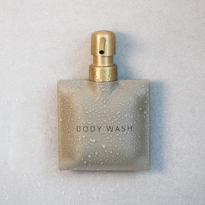 Peut inclure: Un flacon de gel douche carré de couleur beige avec une pompe distributrice en or brossé. La surface du flacon présente une finition texturée et est recouverte de petites gouttelettes d'eau. Les mots BODY WASH sont imprimés au centre.