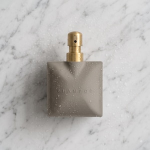 Puede incluir: Un frasco de champú cuadrado de color gris mate con un dosificador de bomba en oro metálico. La palabra SHAMPOO está grabada en la superficie frontal. El exterior del recipiente está cubierto de pequeñas gotas de agua, dándole un aspecto húmedo.