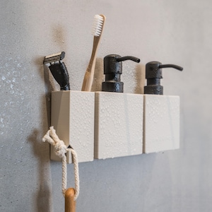 May include: White bathroom organizer with three compartments, holding two black soap dispensers and a toothbrush. A razor and a white rope are hanging from the organizer.