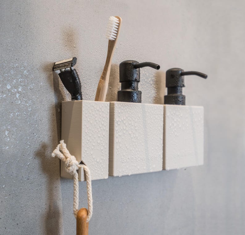 May include: White bathroom organizer with three compartments, holding two black soap dispensers and a toothbrush. A razor and a white rope are hanging from the organizer.