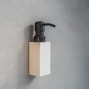 Può includere: Un dispenser di sapone bianco rettangolare con una superficie strutturata. Il dispenser ha una testa della pompa nera strutturata con un beccuccio curvo. Il dispenser è montato su un muro grigio.