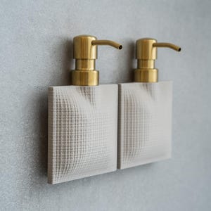 Pode incluir: Dois dispensadores de sabonete combinando com bases quadradas texturizadas em branco sujo e bombas douradas. Os dispensadores são montados em uma parede cinza claro, criando uma estética minimalista e moderna.