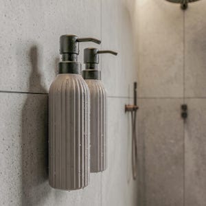 Op de afbeelding: Twee cilindrische zeepdispensers met een verticale geribbelde textuur en een matte lichtgrijze afwerking. Elke dispenser is voorzien van een donkerbrons pompmechanisme aan de bovenkant, met zichtbare waterdruppels op de buitenkant.