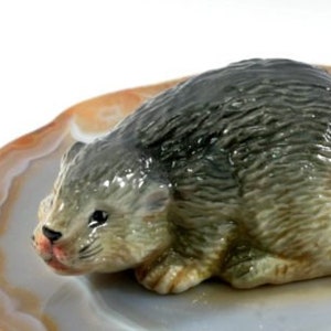 May include: A ceramic beaver figurine, with a grey and brown body and a pale tail. The beaver rests on a polished agate stone, displaying brown, white, and orange bands. The figurine has a glossy finish.