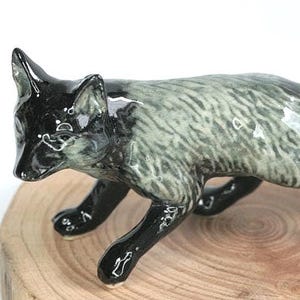 Könnte beinhalten: Eine Keramik-Fuchsfigur in Gehposition. Der Fuchs hat einen glänzenden schwarzen Kopf und Beine mit einem grau-weißen Körper. Der Schwanz ist weiß gespitzt. Die Figur wird auf einer Holzoberfläche präsentiert.