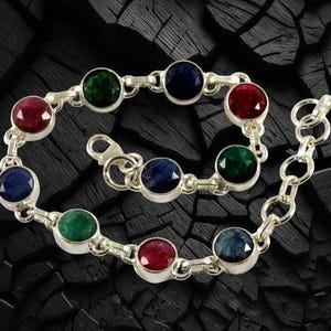 Puede incluir: Pulsera de plata con piedras preciosas redondas en rojo, verde y azul. Las piedras están engastadas en biseles individuales y unidas por una cadena. La pulsera se muestra sobre un fondo oscuro y texturizado.