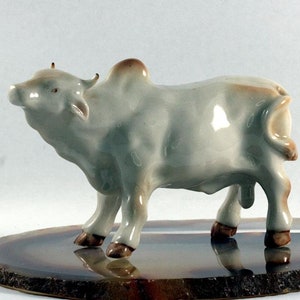 Puede incluir: Una figurita de cerámica blanca de una vaca con detalles marrones. La vaca está de pie sobre una rodaja de ágata redonda, marrón y gris.