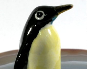 May include: A ceramic penguin figurine with a glossy finish. The penguin has a black head and back, a yellow belly, and black feet. The beak is yellow, and the eyes are white with black pupils. The figurine is standing upright.