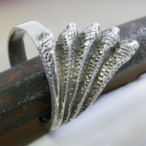 Könnte beinhalten: Ein silberner Ring mit einem einzigartigen Design aus fünf Schlangenköpfen. Der Ring hat ein breites Band, und die Schlangenköpfe sind aufwendig detailliert, was ein auffälliges Schmuckstück ergibt.