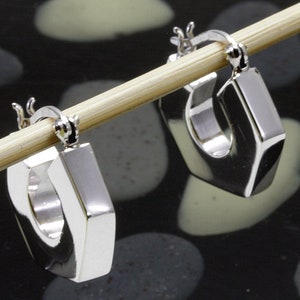 Op de afbeelding: Een paar zilveren creolen met een zeshoekige vorm. De oorbellen hangen aan een houten stok.