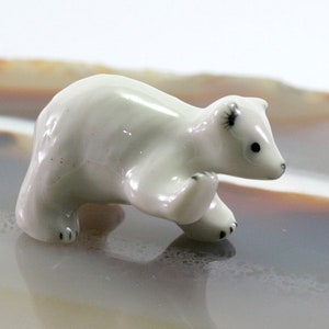 Op de afbeelding: Een kleine, witte keramische ijsbeerfiguur. De beer wordt afgebeeld in een loopstand met zwarte accenten op zijn poten, oren en ogen. Het beeldje heeft een glanzende afwerking en staat tegen een neutrale achtergrond.
